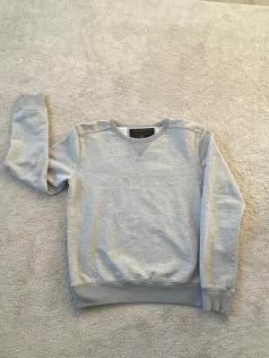 Ljusgrå sweatshirt från Morris - Ljusgrå sweatshirt från Morris med broderad logga och klöverdetalj på bröstet. Tröjan har rund halsringning, ribbade muddar vid ärmslut och nederkant samt en klassisk relaxed fit. Perfekt för en chill och stilren look.