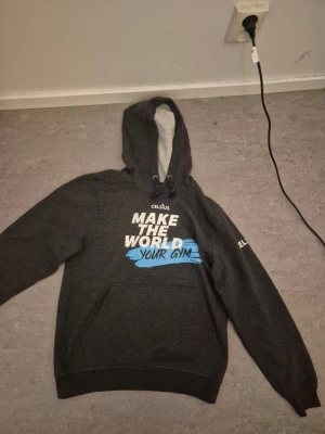 Mörkgrå hoodie från Celsius - Snygg mörkgrå hoodie från Celsius med vit och blå text 'MAKE THE WORLD YOUR GYM' på bröstet och Celsius-logga på ärmen. Hoodien har en stor ficka framtill och justerbar huva med dragsko. Perfekt för dig som gillar sportig och avslappnad stil.