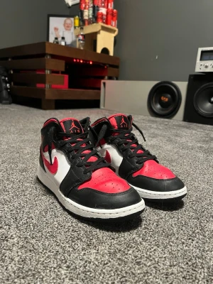 Nike Air Jordan 1 High röd/svart/vit - Säljer ett par Nike Air Jordan 1 High sneakers med klassisk färgblockning i rött, svart och vitt. Skorna har högt skaft, svart snörning och den ikoniska röda swoosh-loggan på sidan. Materialet är skinn och syntet, med rund tå och platt sula.