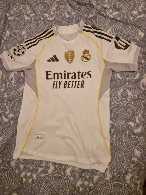 Real Madrid matchtröja Adidas vit - Snygg vit Real Madrid matchtröja från Adidas med gula och grå detaljer. Tröjan har klubbmärke, sponsortryck och Champions League-märke på ärmen. Tillverkad i lätt och ventilerande material, perfekt för fotboll eller träning. Tröjan har en väldigt liten fläck under Adidas märket men går bort efter några tvättar.(Pris går att ändras)