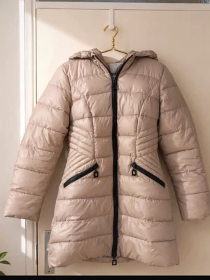 Beige lång dunjacka med huva - Snygg beige dunjacka i längre modell med huva och svarta dragkedjor. Jackan har två snedställda fickor framtill och quiltad design som ger en schysst siluett. Perfekt för kalla vinterdagar och riktigt varm tack vare dunfyllningen.