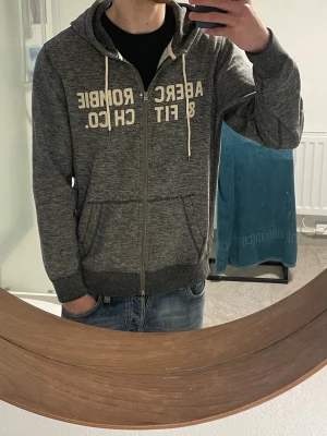 Grå hoodie från Abercrombie & Fitch - Säljer en grå zip-hoodie från Abercrombie & Fitch med vita snören och tryck framtill. Hoodien har två fickor, dragkedja och mjukt material på insidan. Perfekt för chill dagar och enkel att matcha med jeans eller joggers.