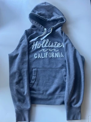 Grå hoodie från Hollister California - Mysig grå hoodie från Hollister California med broderad logga och text framtill. Klassisk känguruficka, huva med vita snören och mjukt material. Perfekt för chill dagar och enkel att matcha med jeans eller joggers.
