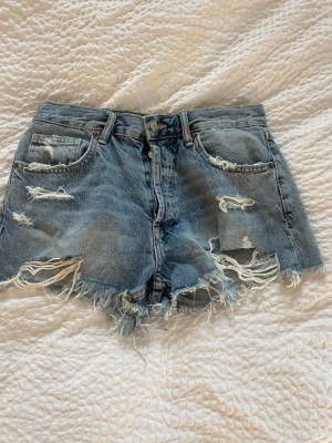 Blå midwaist jeansshorts med fransar - Säljer ett par blå  jeansshorts från Zara med slitna detaljer och råa fransar längs bensluten. Helt oanvända!