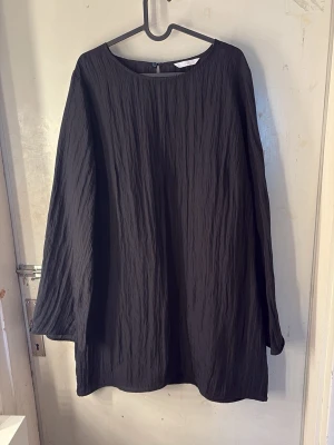 Svart långärmad kortklänning - En svart, enkel och stilren kortklänning med lång ärm och rund halsringning. Klänningen har en lätt strukturerad, skrynklig yta och knäppning bak i nacken. Passar perfekt för dig som gillar minimalistisk och clean stil.