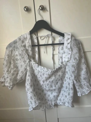 Vit blommig blus med puffärm - Söt vit blus med små blommor och puffiga korta ärmar. Blusen har volangkant vid halsen, smockad struktur och knytband i nacken.