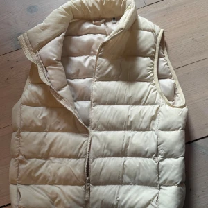 Beige quiltad dunväst med hög krage fr Uniqlo - Stilren beige dunväst med quiltat mönster och hög krage. Västen har dragkedja framtill och är ärmlös, perfekt för lager på lager. Materialet är mjukt och puffigt vilket ger en modern look. Passar till många olika outfits.
