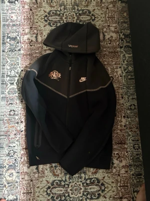 Syna world Nike tech - Säljer en svart hoodie från Nike med hel dragkedja och huva. Framtill finns vita tryck med Nike-logga och ett grafitti-inspirerat motiv samt texten 'LIFE OF SYN' på huvan. Hoodien har kontrastsömmar och sportig vibe. Perfekt för dig som gillar streetwear och snygga detaljer.