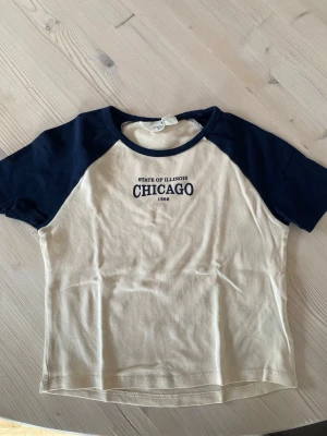 Beige och marinblå Chicago t-shirt H&M - Snygg t-shirt från H&M i beige med marinblå ärmar och krage. Tryck med texten 'State of Illinois Chicago 1992' på bröstet. Klassisk raglanmodell med korta ärmar, perfekt för en avslappnad look. Köparen står för frakt.