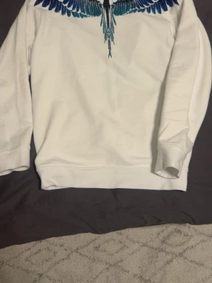 Vit sweatshirt med blått vingmönster - Säljer en vit sweatshirt från Marcelo Burlon med ett ikoniskt blått och svart vingmönster runt halsen. Tröjan har rund halsringning, långa ärmar och är gjord i mjukt bomullsmaterial. Perfekt för dig som vill sticka ut med en unik design.