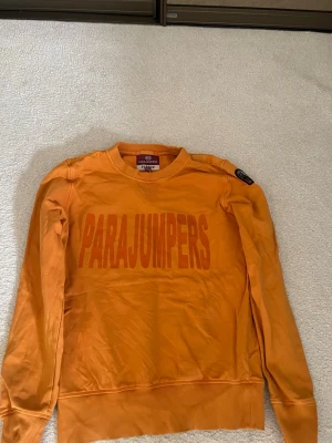 Orange sweatshirt från Parajumpers - Snygg orange sweatshirt från Parajumpers med stort tryck på bröstet och rund hals. Tröjan har långa ärmar, ribbade muddar och en patch på axeln. Perfekt för dig som gillar streetwear och vill sticka ut lite extra.