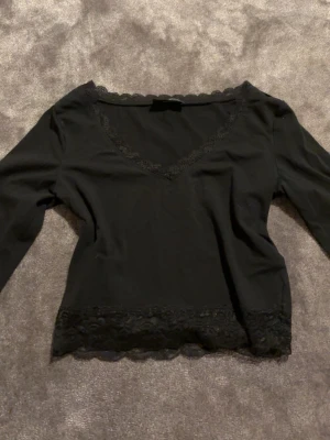 Svart topp med spetsdetaljer - Svart långärmad topp med v-ringning och spetsdetaljer längs halsringningen och nederkanten. Tunn och mjuk, perfekt att styla med jeans eller kjol. Enkel men ändå elegant design som ger en snygg touch till din outfit.