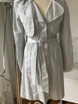Ljusgrå/silver trenchcoat med knytskärp - Snygg ljusgrå /silver trenchcoat med bred krage, knytskärp i midjan och smockdetaljer. Jackan har stora slag, knappstängning framtill och två fickor. Tillverkad i ett tunt, mjukt tyg som ger en avslappnad men stilren look.