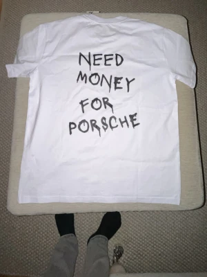 Vit T-shirt med text ”need money for Porsche” - Säljer en vit T-shirt från med svart text på ryggen där det står 'NEED MONEY FOR POR$CHE' i handskriven stil. Klassisk passform med rund hals och korta ärmar. Tillverkad i mjuk bomull och har en tagg kvar i nacken.
