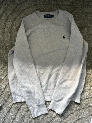 Grå sweatshirt från Polo Ralph Lauren - Klassisk ljusgrå sweatshirt från Polo Ralph Lauren med rund halsringning och den ikoniska mörkblå logobroderingen på bröstet. Tröjan har ribbade muddar vid ärmslut och nederkant, perfekt för en avslappnad och stilren look.