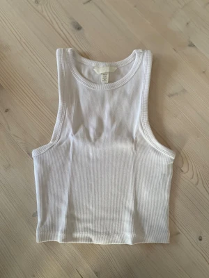 Vit ribbad linnetopp från H&M - Säljer en vit ribbad linnetopp från H&M i storlek XS. Toppen är croppad och ärmlös med rund halsringning och stretchigt material. Köparen står för frakt.