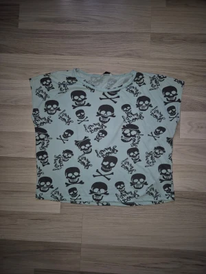 Turkos topp med dödskallemönster - Cool turkos topp med svart dödskallemönster och lös passform. Perfekt för dig som gillar edgy stil och vill sticka ut. Toppen är ärmlös och har rund halsringning. Materialet känns mjukt och skönt mot huden. Den har 2 små hål, se bild. 