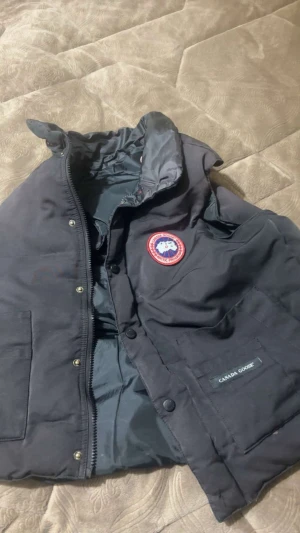 Svart vest från Canada Goose - Säljer en svart dunjacka från Canada Goose med klassisk röd logotyp på bröstet och diskret varumärkesmärke på fickan. Jackan har hög krage, dragkedja och tryckknappar framtill samt stora fickor. Perfekt för kalla vinterdagar.     Funkar ej att stänga