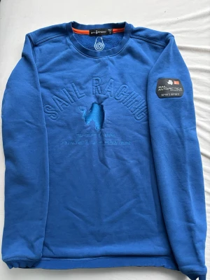 Blå Sail Racing sweatshirt - Blå sweatshirt från Sail Racing med broderad logga och text på bröstet samt patch på ärmen med 'Sail Antarctica Expedition'. Rund halsringning och ribbade muddar. Perfekt för dig som gillar sportig och stilren design.