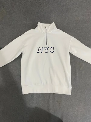 Vit halfzip hoodie med NYC-tryck - Snygg vit hoodie med halv dragkedja och hög krage. Framtill står det 'NYC' i svart text. Hoodien har långa ärmar och ribbade muddar. Perfekt för en avslappnad streetstyle-look. Materialet känns mjukt och bekvämt, troligen bomullsblandning.
