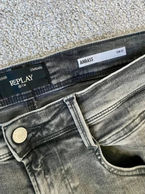 Replay Anbass grå slim fit jeans - Snygga grå jeans från Replay, modell Anbass med slim fit passform. Jeansen har klassisk femficksdesign, silverfärgad knapp och diskreta slitningar för en cool look. Perfekta till streetwear eller en avslappnad stil. Använda en gång 