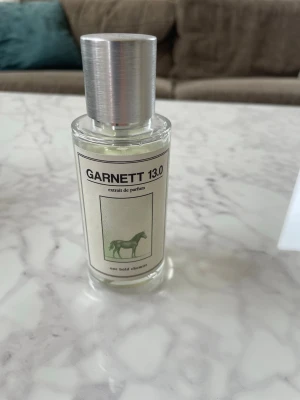6 parfymer  - Två parfymer: Garnett 13.0 extrakt de parfum i en genomskinlig glasflaska med silverfärgat lock och minimalistisk etikett med hästmotiv. Afnan 9PM Rebel i en rektangulär flaska med spegelblank framsida, svart text och rund silverkula som lock, levereras i färgstark förpackning.