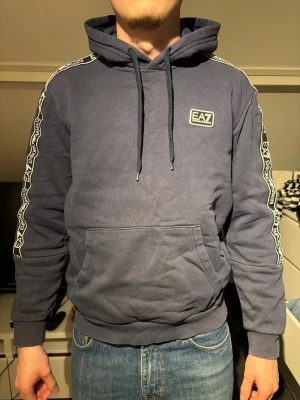 Mörkblå hoodie från EA7 Emporio Armani - Snygg mörkblå hoodie från EA7 Emporio Armani med huva och dragsnören. Har vita detaljer längs ärmarna och en EA7-logga på bröstet. Känguruficka framtill och ribbade muddar vid ärmslut och midja. Perfekt för en avslappnad och trendig look.