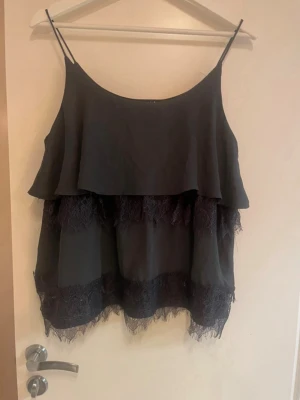 Mörkgrå linnetopp med spetsdetaljer - Snygg mörkgrå linnetopp med tunna axelband och lager av spetsdetaljer. Toppen har en lös passform och volangkant som ger en luftig känsla. Perfekt att styla med jeans eller kjol för en trendig look.