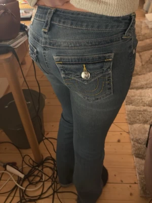 Blå bootcut jeans från True Religion - Säljer ett par blå bootcut jeans från True Religion Jeansen har låg midja och detaljerade bakfickor med gul söm och knapp. Klassisk denim-look som passar till allt. Knappt använda!!