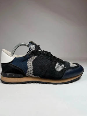 Valentino camo sneakers i mesh och mocka - Snygga sneakers från Valentino med camomönster i svart, grått och blått. Ovandelen är i mesh och mocka med vita och mörkblå detaljer samt en vit hälkappa. Gummisula med nitar på hälen ger en unik touch. Perfekta för dig som vill sticka ut med stil. Storlek 42 men passar 43