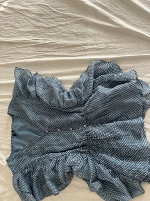 Blå blus med volanger - Trendig blå blus med små vita mönster och volangdetaljer. Blusen har knappar framtill och en luftig, flowy passform som ger en romantisk vibe. Perfekt för dig som gillar en feminin och avslappnad stil.