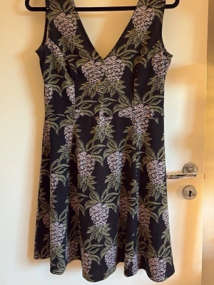 Svart blommig klänning med v-ringning - Snygg svart klänning med v-ringning och ärmlös design. Klänningen har ett blommigt mönster i lila och grönt som ger en fräsch känsla. Passar perfekt till sommaren och har en lätt utställd kjol för en bekväm passform.