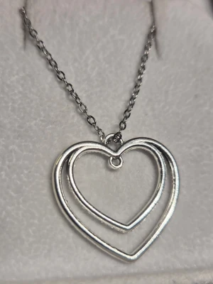 Silverfärgat halsband med dubbla hjärtan - Snyggt halsband i silverfärg med två öppna hjärtan som hänger i varandra. Kedjan är tunn och smidig, och hjärtpännet ger en romantisk touch till din stil. Perfekt för dig som gillar minimalistiska smycken med en söt detalj. Kedjan är av rostfritt stål, 45 cm. 