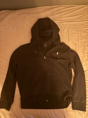 Svart hoodie från Polo Ralph Lauren - Svart hoodie med dragkedja från Polo Ralph Lauren. Klassisk design med huva och dragsko, två stora fickor framtill och den ikoniska vita logotypen broderad på bröstet. Tillverkad i mjukt bomullsmaterial som är skönt att bära.