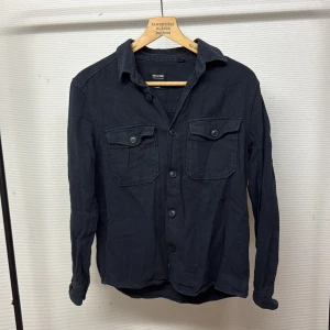 Mörkblå overshirt från Only & Sons - Snygg mörkblå overshirt från Only & Sons med två bröstfickor med lock och knappar. Skjortan har klassisk krage, långa ärmar och är tillverkad i mjuk bomull. Perfekt att bära öppen över en t-shirt för en avslappnad look.