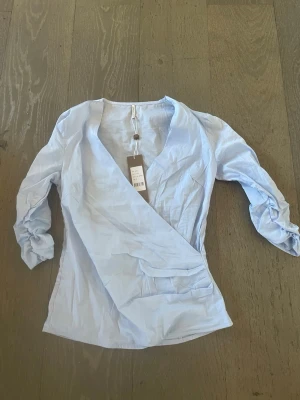 Ljusblå omlottblus med trekvartsärm - Snygg ljusblå omlottblus med djup v-ringning och trekvartsärmar. Blusen har en feminin passform och knytdetalj i sidan. Tillverkad i ett lätt och luftigt material som känns skönt mot huden. Perfekt att styla med jeans eller kjol.