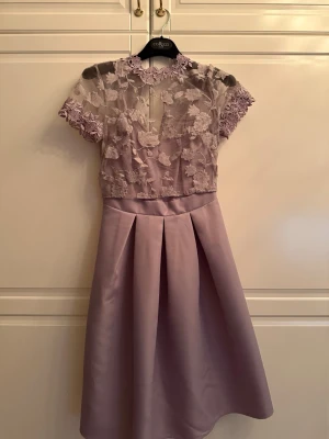 Ljuslila klänning med spetsdetaljer - Supersnygg ljuslila klänning med korta ärmar och transparent överdel i spets med blommönster. Kjolen är i satinliknande material med veck framtill. Hög hals med dekorativa spetskanter och dragkedja bak.