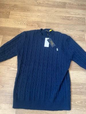 Mörkblå kabelstickad tröja Polo Ralph Lauren - Snygg mörkblå kabelstickad tröja från Polo Ralph Lauren med klassisk rund halsringning och den ikoniska vita logon broderad på bröstet. Tröjan har långa ärmar och ribbade muddar vid ärmslut och nederkant. Perfekt för lager på lager och stilren vardagslook.
