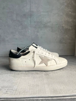 Golden Goose vita sneakers med stjärna - Snygga vita sneakers från Golden Goose med grå mockastjärna på sidan och svart hälparti. Skorna har klassisk låg profil, vita skosnören och är tillverkade i läder med detaljer i mocka. Perfekta för en trendig och avslappnad stil.