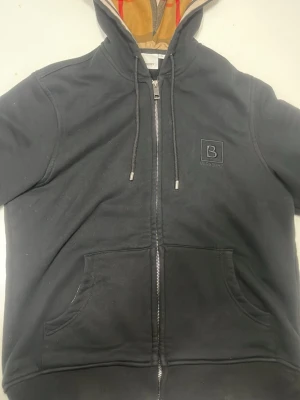 Svart hoodie från Burberry med dragkedja - Svart hoodie från Burberry med dragkedja och klassiskt rutigt foder i huvan. Broderad B-logga på bröstet, två stora fickor framtill och justerbara snören i huvan. Perfekt för dig som gillar stilrena och exklusiva plagg.