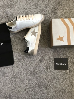 Golden Goose vita sneakers med stjärna - Golden Goose sneakers i vitt läder med grå stjärna på sidan, beige skosnören och svart detalj på hälen. Klassisk låg modell med slitna detaljer för en cool och avslappnad look. Kommer med originalkartong och dustbag. (Rep)