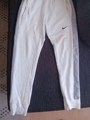 Nike mjukisbyxor - Säljer ett par vita mjukisbyxor från Nike med grå sidopaneler och diskret Nike-logga på vänster lår. Byxorna har resår i midjan, fickor på sidorna och muddar vid bensluten. Perfekta för chill dagar eller träning.