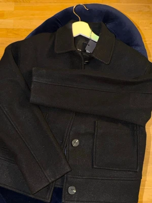 Svart ulljacka från lager 157 - Stilren svart jacka från Topman i ullmaterial. Jackan har klassisk krage, stora framfickor och stängs med knappar framtill. Perfekt för dig som gillar minimalistisk och clean stil. Ordnarie pris 400kr 