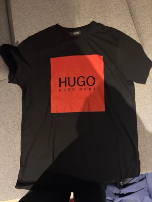 Svart t-shirt från Hugo Boss - Snygg svart t-shirt från Hugo Boss med stort rött tryck framtill där det står HUGO. Klassisk passform och rund hals. Tillverkad i mjuk bomull som känns skön mot huden. Perfekt för en stilren och modern look.