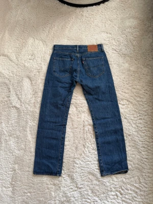 Levi's raka blå jeans 501 - Klassiska blå jeans från Levi's med rak passform och fem fickor. Jeansen har en mörkblå tvätt, synliga sömmar och läderpatch bak i midjan. Tillverkade i slitstark denim, perfekta för en avslappnad och tidlös stil.