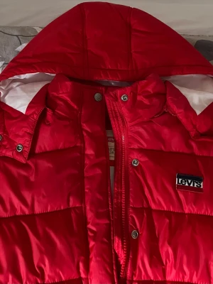 Röd pufferjacka från Levi's - Snygg röd pufferjacka från Levi's med huva och quiltad design. Jackan har dragkedja, tryckknappar och ett tydligt Levi's-märke på bröstet. Insidan är vit och materialet är glansigt syntet. Perfekt för kalla vinterdagar.