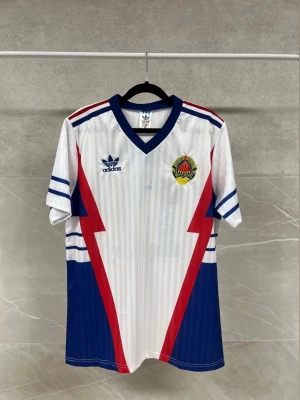 Jugoslavien retro fotbollströja Adidas - Retro fotbollströja från Adidas i vitt med blå och röda blixtdetaljer, blå krage och ärmslut. Tryck med namn 'MIHAJLOVIĆ' och nummer 11 på ryggen. Broderad klubbmärke på bröstet och klassiska Adidas-loggan. Perfekt för dig som gillar vintage sportstil.