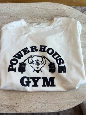 Vit Powerhouse Gym t-shirt - Säljer en vit t-shirt från Powerhouse Gym med stor svart logga och illustration framtill. T-shirten är i 100% polyester och har klassisk rund hals och korta ärmar. Perfekt för dig som gillar gym och träning.
