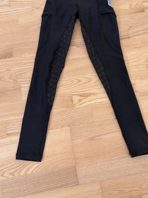 Svarta sportiga ridleggings från Maya Delores med mönster för ett bra grepp.  - Svarta ridleggings från Maya Delores med sportig look och diskret mönster längs insidan av benen. Tight passform och stretchigt material som passar perfekt till träning. Snygga sömmar och hög midja för extra komfort. Ett ihopsytt hål längst ner vid vänster ben.