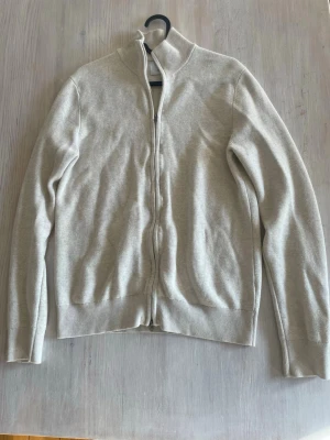 Ziptröja från Bläck - Stilren beige/vit ziptröja från Bläck med hög krage och hel dragkedja framtill. Tröjan har ribbade muddar vid ärmslut och nederkant, vilket ger en snygg passform. Perfekt att slänga på sig över en t-shirt för en clean look.